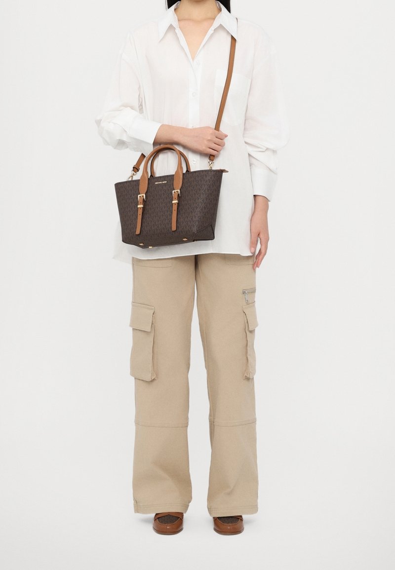 Femme portant un chemisier blanc et un pantalon cargo beige tenant un sac à main de créateur marron avec des sangles camel et une bandoulière.