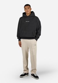 Svart hettegenser med frontlomme og "PEGADOR"-logo, sammen med beige bukser med tydelig piping og brodert logo. Sorte sneakers.