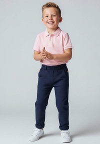 Młody chłopiec uśmiechający się, ubrany w różową koszulkę polo, granatowe spodnie i białe trampki, stoi na tle jednolitego jasnoszarego tła.