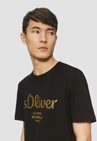 Zwarte katoenen T-shirt met een gouden logo met de tekst "s.Oliver Elevated Essentials" in een ontspannen pasvorm en ronde hals.