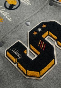 Szary wełniany bomber z ciemnoniebieską i żółtą filcową literą "S" z wyhaftowanymi akcentami, czarnymi zapięciami na zatrzaski oraz ozdobnymi naszywkami.