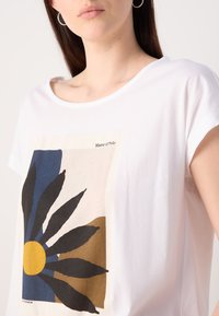 T-shirt en coton blanc avec un grand graphique d'une fleur aux pétales noirs, un centre jaune et des formes marron et bleu en arrière-plan.