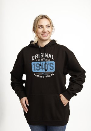 Femme souriante en sweat à capuche noir avec le texte "Original and Unrestored 1940's Vintage Goods", debout les mains dans les poches, regardant à droite.