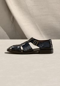 Μαύρο ανδρικό δερμάτινο σανδάλι με υφή κροκοδείλου, με λουράκι με αγκράφα και ανοιχτή μύτη, παρουσιασμένο πάνω σε φωτεινή επιφάνεια.