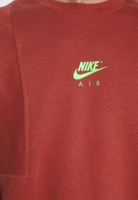 Röd sweatshirt i mjukt material med en grön Nike-logotyp ovanför ordet "AIR." Enkel rund halsringning med synliga sömmdetaljer.