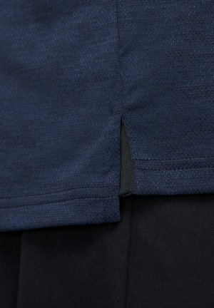 Mörkblå t-shirt i texturerat tyg, med sidoklyftor och kontrasterande svart söm längs fållen. Designen är minimalistisk och strömlinjeformad.