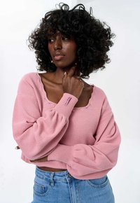 Roze gebreide trui met een textuurpatroon, wijde mouwen en een gegolfde halslijn. Gecombineerd met lichte blauwe spijkerbroek.