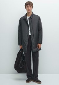 Schwarze gepolsterte Jacke mit Reverskragen, kombiniert mit dunklen Hosen und einer schwarzen Wildleder-Tasche. Das Modell trägt ein weißes Oberteil und Loafers.