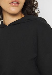 Felpa nera in cotone con cappuccio regolabile, caratterizzata da una consistenza morbida e una vestibilità rilassata. Primo piano della zona del collo e della spalla.