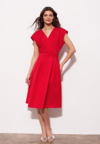 Czerwona sukienka do kolan z krótkimi rękawami, dekoltem w kształcie litery V i dwoma guzikami w talii. Gładki materiał i zwiewna spódnica. Srebrne sandały na otwartym palcu.