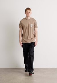 Brun kortermet t-skjorte med brystlomme, kombinert med svarte bukser. Stoffet ser mykt ut og har en avslappet design.