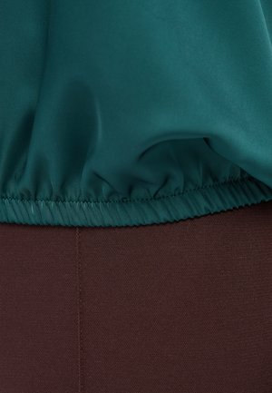 Teal stof met een geplooide rand over een gladde donkerbruine stof gedrapeerd, waarbij de stiksels en de textuurcontrasten tussen de materialen zichtbaar zijn.