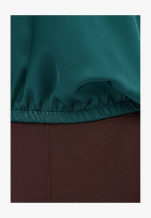 Teal stof met een geplooide rand over een gladde donkerbruine stof gedrapeerd, waarbij de stiksels en de textuurcontrasten tussen de materialen zichtbaar zijn.