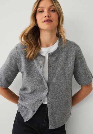 Woman with blonde hair wearing a grey short-sleeve buttoned cardigan over a white top and black pants, standing against a plain wall.