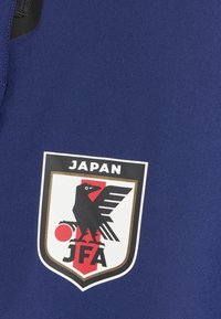 Camisola desportiva azul com o logotipo da Associação de Futebol do Japão, com um pássaro preto, detalhe vermelho e contorno branco. Texto "JAPAN" acima do emblema.