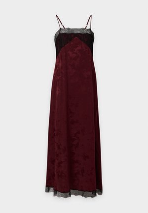 Femme aux cheveux bouclés portant une robe midi bordeaux sans manches avec une bordure en dentelle noire et des talons hauts noirs, debout devant un fond uni.