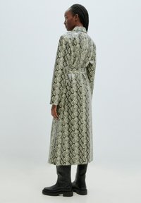 Lange slangenprintjas in tinten van groen en zwart, met een ceintuur in de taille, kraag en een gladde, glanzende textuur. Zwarte kniehoge laarzen.