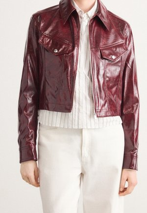 Woman wearing a cropped dark red leather jacket with flap pockets over a white pinstriped button-up shirt and white pants.