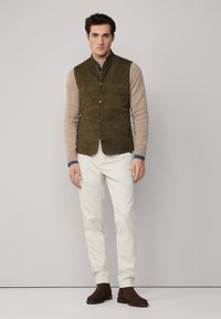 Zelená semišová vesta přes béžovou košili s dlouhým rukávem, doplněná o bílé kalhoty a hnědé kotníkové boty. Minimalistický design s předním zapínáním na knoflíky.