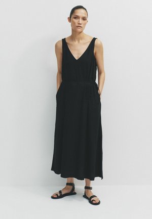 Nő fehér fal előtt áll, fekete ujjatlan midi ruhát visel, amely zsebekkel rendelkezik, valamint fekete bokapántos szandált.