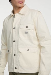 Cremfarbene Baumwolljacke mit einem Kragen, Frontknöpfen, mehreren Taschen und einem kleinen Markenschild. Verfügt über silberfarbene Hardware-Details.