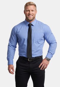 Blonder Mann lächelnd, trägt ein blaues Hemd, schwarze Krawatte, schwarze Hose und eine Armbanduhr, steht mit einer Hand in der Hüfte.