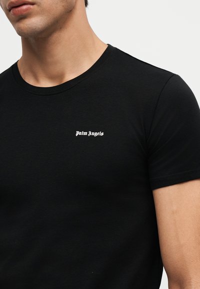 Άνδρας που φοράει απλό μαύρο κοντομάνικο T-shirt με μικρό λευκό κείμενο "Palm Angels" στο αριστερό στήθος.