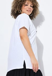T-shirt en coton blanc à manches courtes, avec des fentes sur les côtés. Le mannequin pose avec des cheveux bouclés, portant le t-shirt par-dessus une jupe noire.