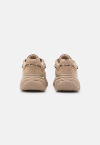 Beige athletic shoes with a textured upper, rounded toe, and thick rubber soles. Features side branding and contrasting heel accents.