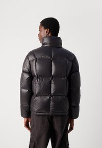 Juoda pūkinė striukė su aukštu apykakle, kvadratinio siuvinėjimo raštu ir blizgiu paviršiumi. Iš nugaros pusės matosi laisvas siluetas ir elastingi rankogaliai.