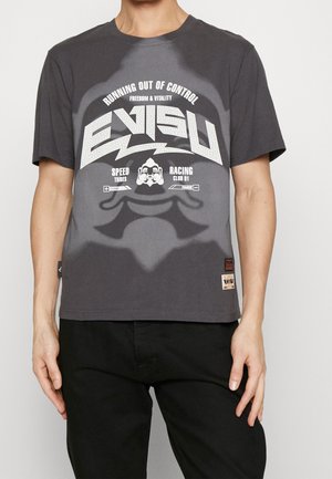 Person iført en mørkegrå Evisu t-shirt med hvid grafisk tekst og et stiliseret ansigtsdesign foran, kombineret med sorte bukser.