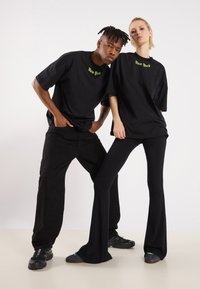 Tricouri negre supradimensionate cu "New York" în verde neon, asortate cu pantaloni negri largi și colanți ajustați din tricot, purtate cu pantofi asortați diferit.
