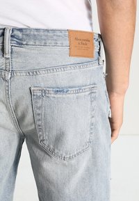 Vaaleansininen farkkuhousu henkilöllä, jossa näkyy takatasku ja ruskea Abercrombie & Fitch -nahkaleima.