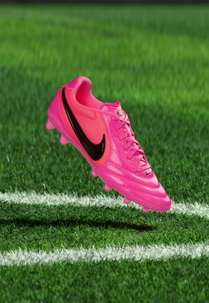 Adidaș de fotbal Nike roz aprins cu swoosh negru, plutind deasupra ierbii verzi lângă liniile albe ale terenului.