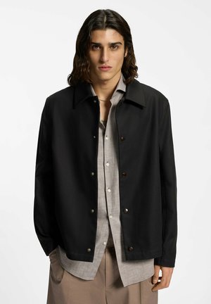 Giovane con capelli alla lunghezza della spalla che indossa una giacca nera abbottonata sopra una camicia grigio chiaro e pantaloni beige su uno sfondo bianco.