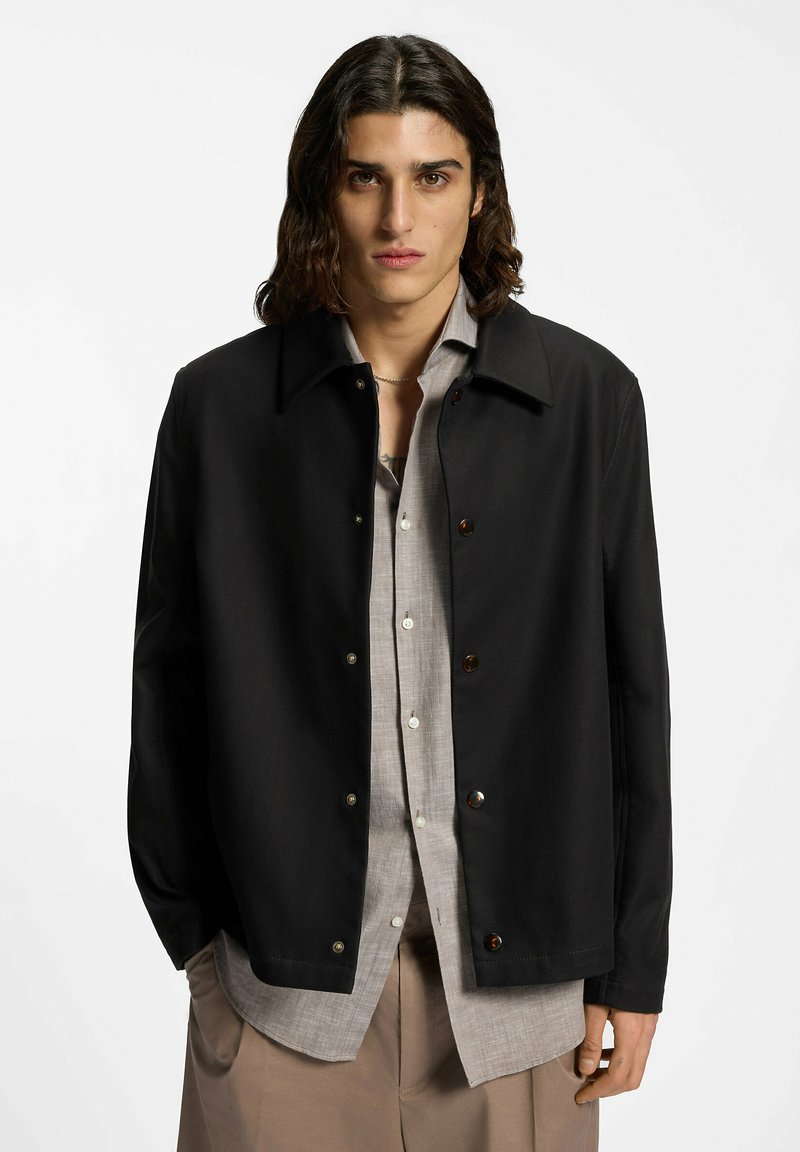 Jeune personne aux cheveux mi-longs portant une veste noire boutonnée sur une chemise gris clair et un pantalon beige, sur fond blanc.