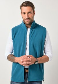 Teal Fleece-Weste mit hohem Kragen, Reißverschluss und seitlichen Taschen, getragen über einem weißen Langarmshirt. Braune Uhr als Akzent.