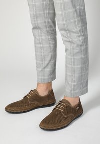 Braune durchbrochene Leder Schuhe mit beigen Schnürsenkeln und einer schwarzen Gummisohle, getragen mit hellgrauen Karohosen.
