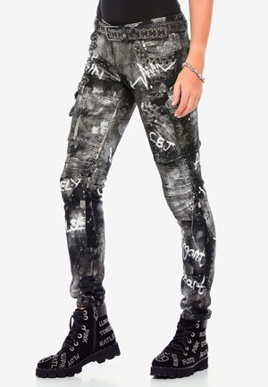 Femme portant un pantalon cargo imprimé graffiti noir et blanc, une ceinture cloutée, un bracelet argenté et des bottes noires à lacets cloutées.