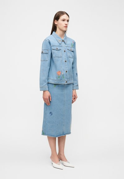 Kvinde iført lyseblå denimjakke og matchende midinederdel med broderede blomsterdesigns, iført hvide spidse hæle, stående foran en hvid baggrund.