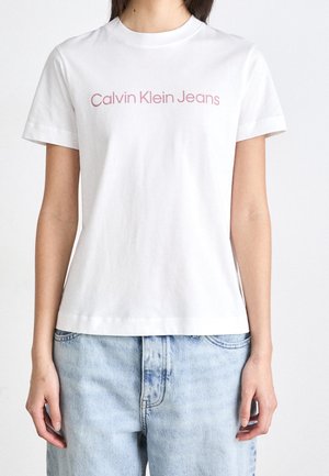 Person iført en hvid T-shirt med korte ærmer med teksten "Calvin Klein Jeans" i lyserød, kombineret med lyseblå denimjeans.