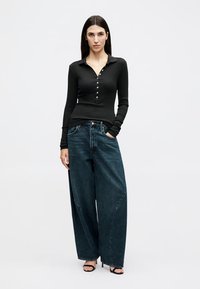 Femme debout portant un haut noir à manches longues boutonné, un jean large bleu foncé et des talons ouverts noirs sur un fond uni.