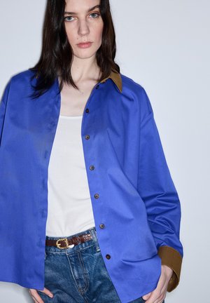 Młoda kobieta o długich ciemnych włosach, ubrana w niebieską koszulę na guziki z brązowym kołnierzykiem i mankietami, białą koszulkę na ramiączkach oraz dżinsy z brązowym paskiem.