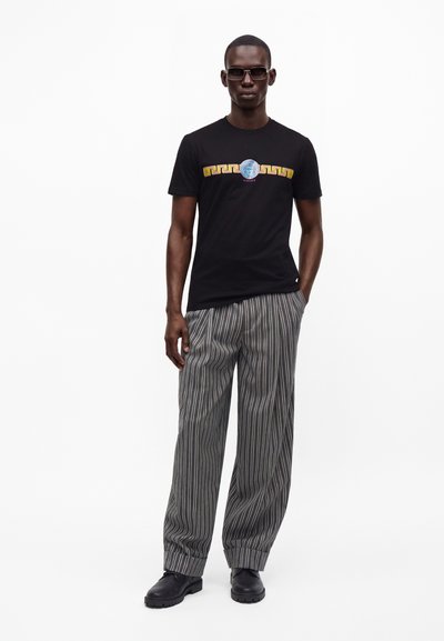 Άνδρας που φοράει μαύρο t-shirt Versace με το λογότυπο Medusa, γκρι ριγωτό παντελόνι με φαρδιά μπατζάκια, μαύρα παπούτσια και γυαλιά ηλίου σε λευκό φόντο.