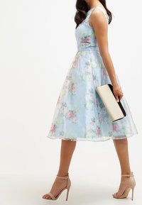 Vaaleansininen kukkakuosinen mekko, jossa on istuva yläosa ja levenevä helma. Mekossa on hienovaraiset ruusukuvioinnit ja läpikuultava päällyskerros. Yhdistetty mustavalkoiseen pikkulaukkuun.