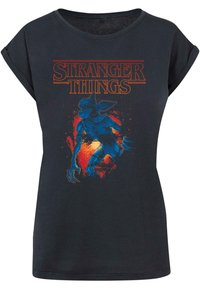 Fekete pamut póló, amely kék lény grafikai elemmel rendelkezik, piros és narancssárga akcentusokkal, "Stranger Things" felirattal, vastag narancssárga betűkkel.