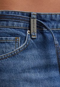 Izbliza plavi traperice s vidljivim prednjim džepom, petljom za pojas i metalnom oznakom s natpisom "2Y STUDIOS" na pozadini kože.