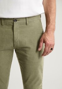 Olivené kordové nohavice so slim fit, s gombíkovým zapínaním a jemnou textúrou, v kombinácii s bielou košeľou. Ruka spočíva vedľa nohavíc.