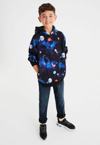 Sudadera con capucha negra con un diseño de temática espacial que incluye planetas de colores y estrellas. Combinada con jeans oscuros y zapatillas deportivas.