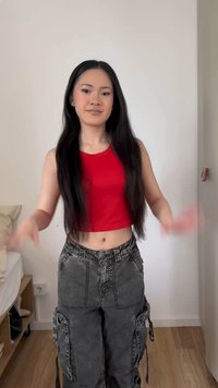 Μια νεαρή γυναίκα με μακριά μαύρα μαλλιά που φοράει ένα κόκκινο crop top και σκούρα γκρι παντελόνια cargo, στέκεται σε ένα δωμάτιο με ξύλινο πάτωμα και λευκούς τοίχους.